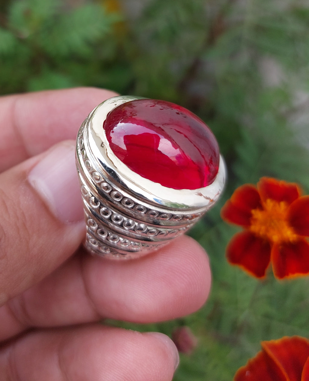 A silver-toned signet ring with a large, polished red cabochon gemstone, held in a person's hand against a blurred background of green foliage and orange flowers.