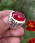 A silver-toned signet ring with a large, polished red cabochon gemstone, held in a person's hand against a blurred background of green foliage and orange flowers.