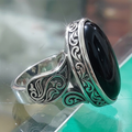 An ornate sterling silver ring with a large oval black onyx cabochon, featuring intricate scrollwork engraving on the band and shoulders, resting on a reflective glass surface.
