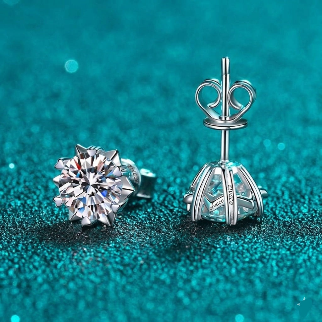 A pair of silver stud earrings displayed on a sparkling teal background. One earring is shown from the front, featuring a brilliant round-cut clear gemstone in a prong setting.