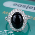 A silver-tone halo ring featuring a large oval black onyx cabochon and surrounded by clear crystals, displayed on a green reflective surface with partial Oakley branding visible.