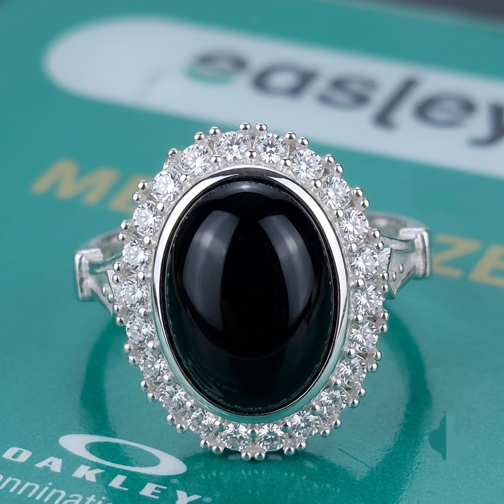 A silver-tone halo ring featuring a large oval black onyx cabochon and surrounded by clear crystals, displayed on a green reflective surface with partial Oakley branding visible.